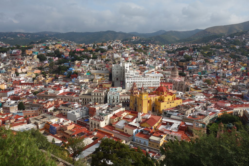In this view from a hilltop overlook, Guanajuato City sprawls out over rolling hills. The main church is prominent in the foreground, dark yellow with two spires in front and a red dome in back. Green, forested hills meet the cloudy horizon beyond the city in the background.