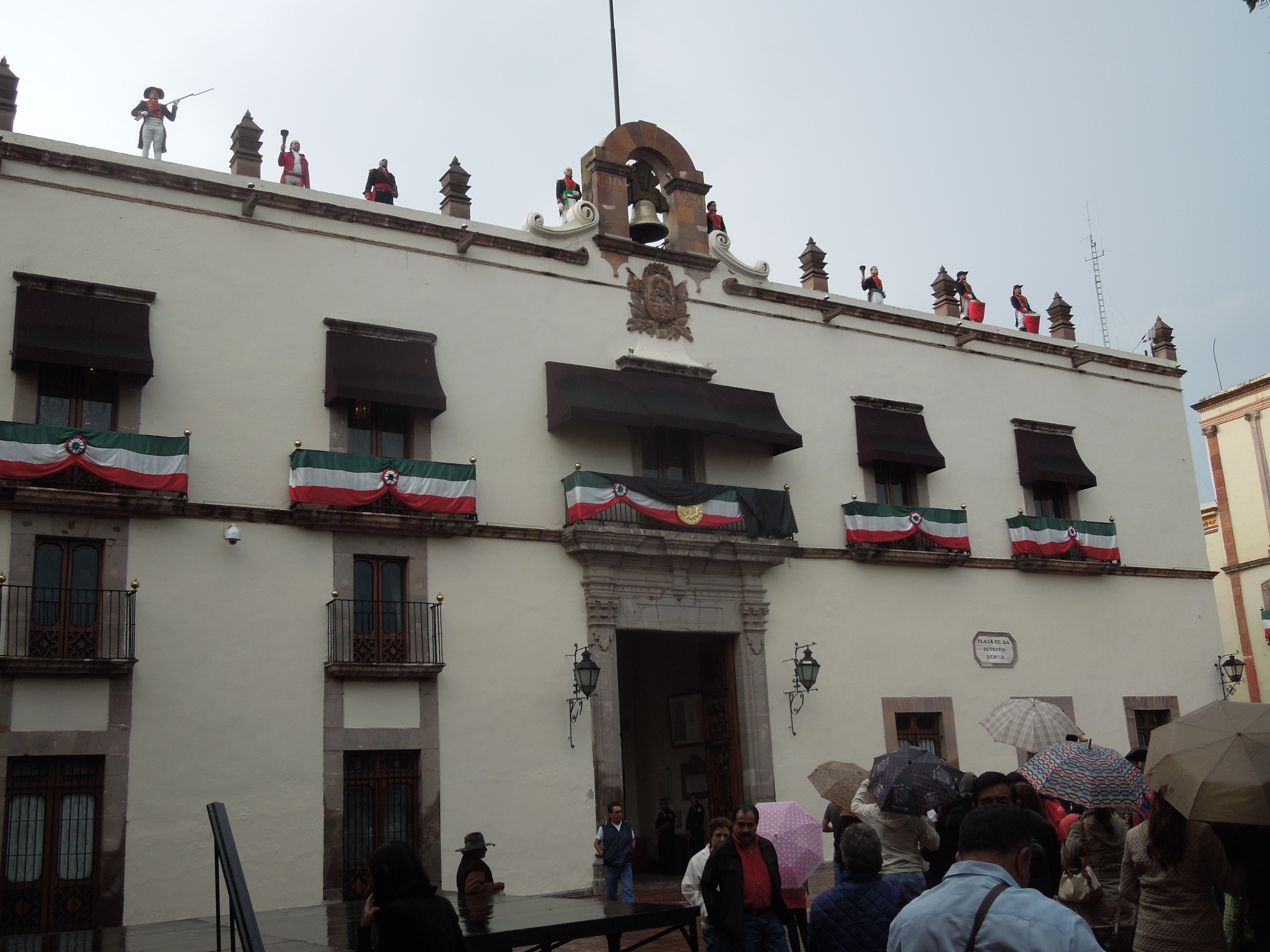 An old government building has balconies adorned with colors of the Mexican flag. A large bell is on the rooftop. Actors stand on top of the building, dressed in contemporary clothing as soldiers, priests, and government figures. 