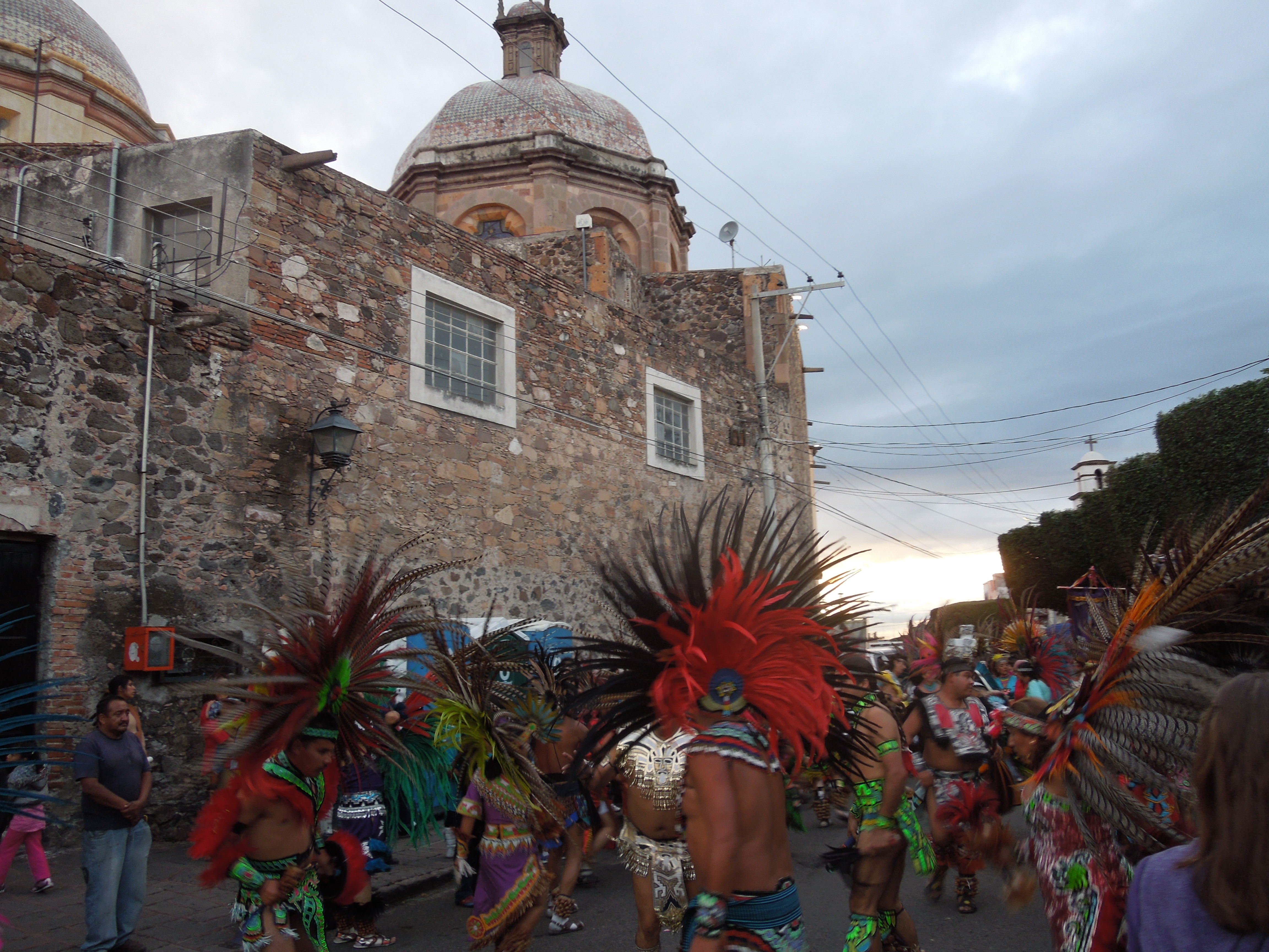 Energetic drummers and dancers in Aztec-style clothing wear headdresses with long feathers radiating outward in all directions. They gather near the old, stone walls of the temple of Santa Cruz. A tower with a dome can be seen on top of the temple. 