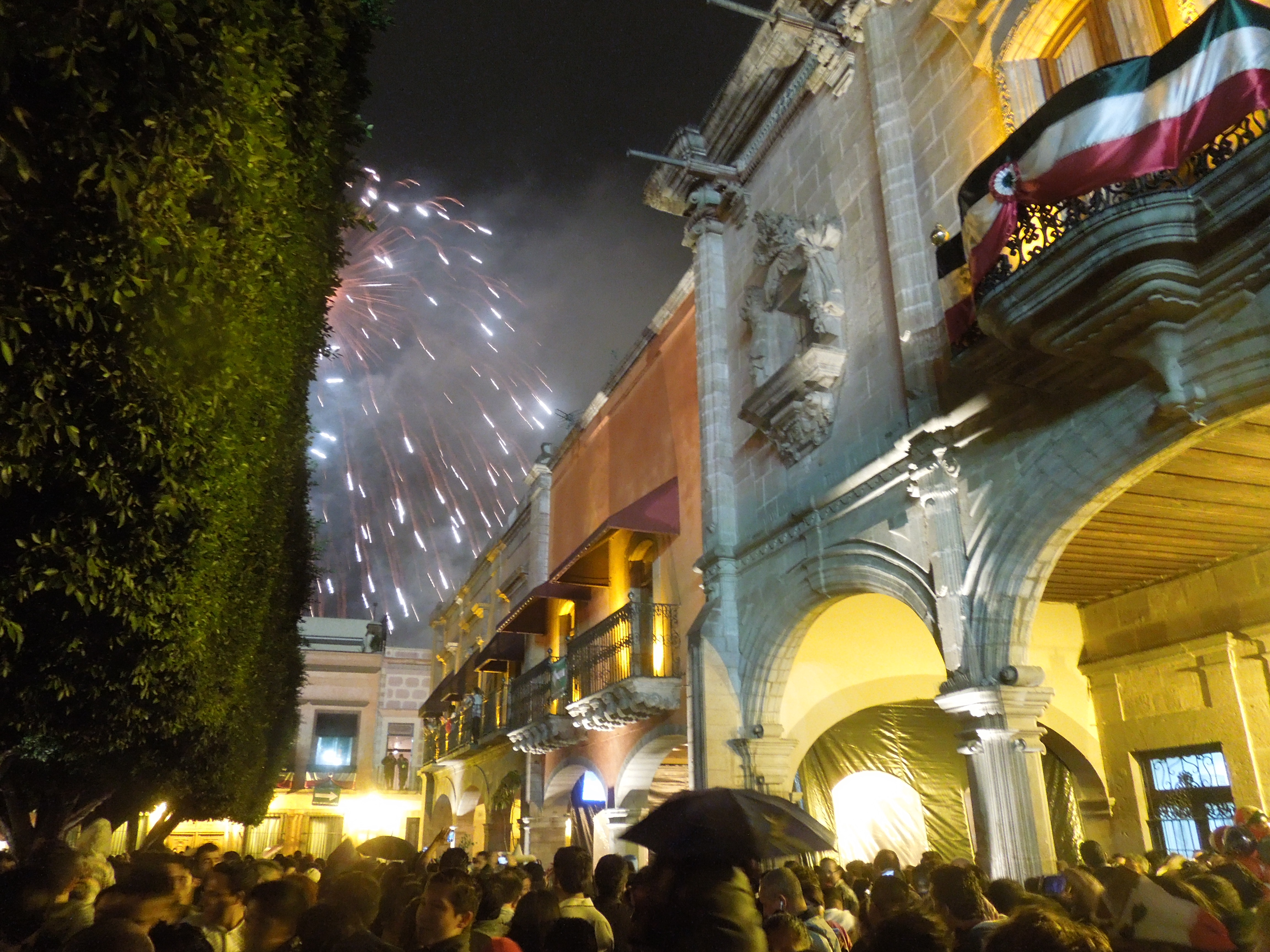Fireworks explode over the Plaza de Armas in Querétaro just moments after the Grito. The fireworks can be seen in the open sky between trees on the left and government buildings adorned with Mexican colors on the right.