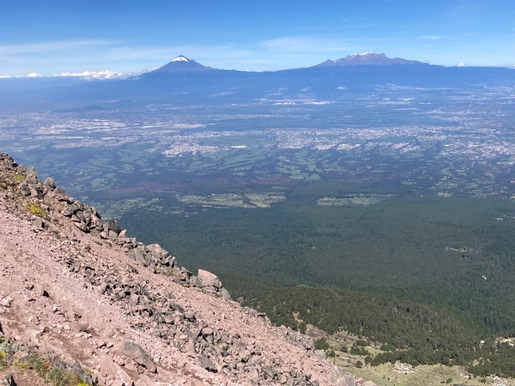 The snow-capped Popocatépetl and Iztaccíhuatl can be seen on the horizon. The rocky slope of La Malinche is in the foreground with dense forests further down, and some cities in the distance. 
